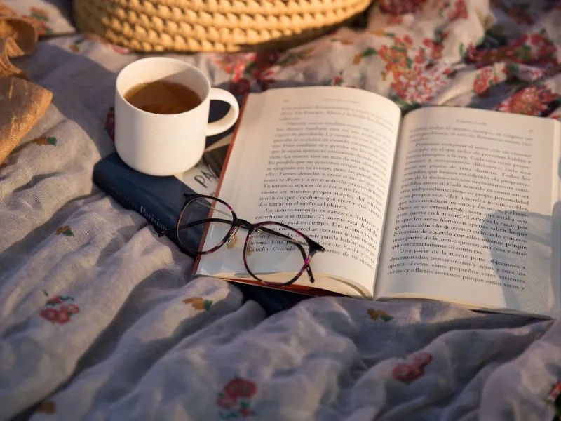An open book, glasses and a cup of tea on a floral blanket