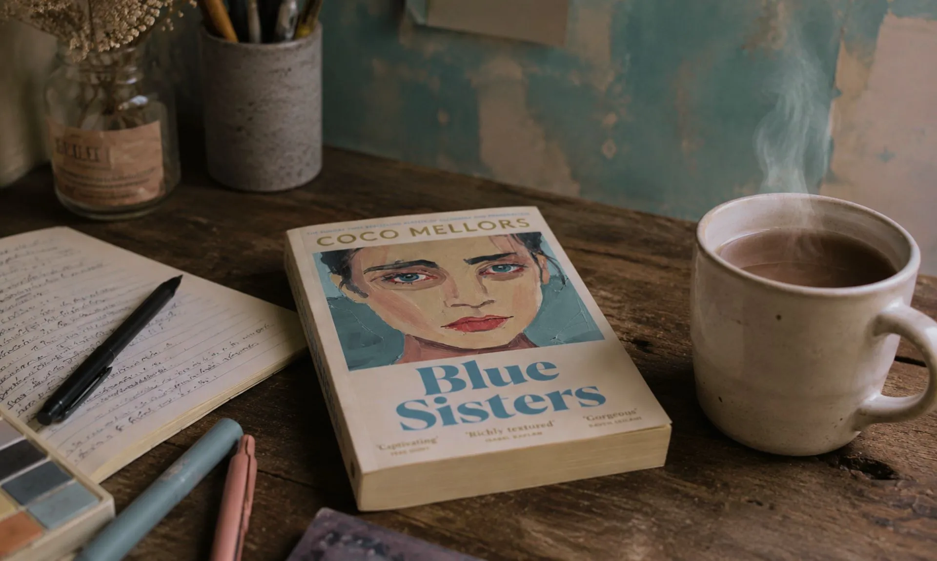 Blue Sisters by Coco Mellors book on a table with writing supplies and a cup of coffee