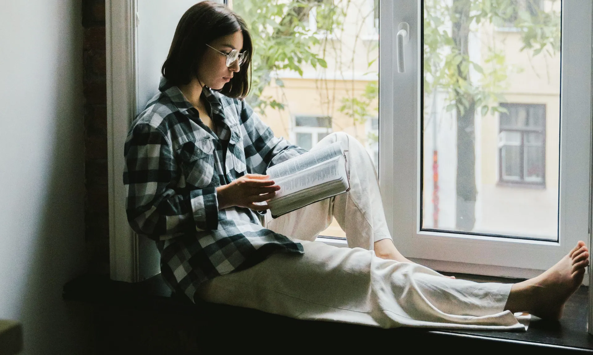a girl sitting near a window reading her book calmly