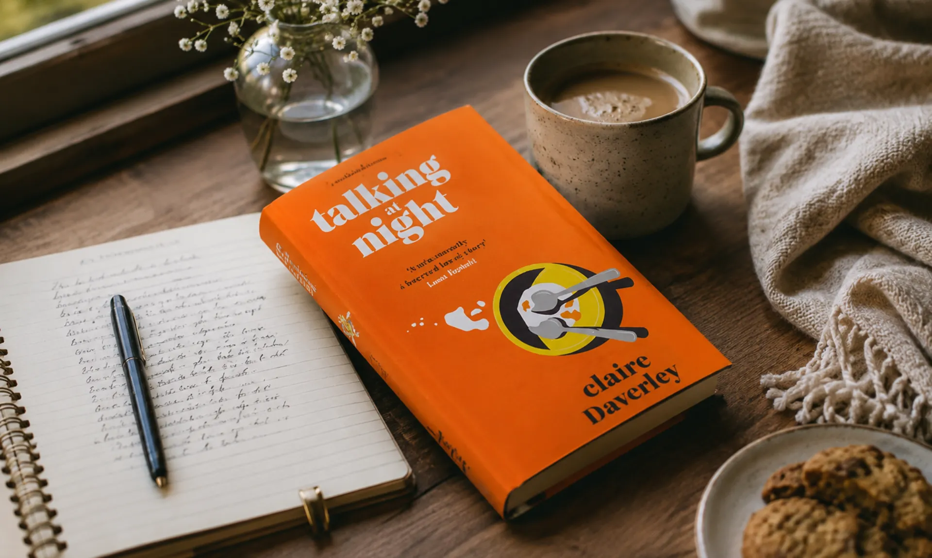 Feature image of Talking at Night by Claire Daverley resting on a wooden table beside a cup of coffee, an open notebook, fresh flowers, cookies, and a cozy blanket, creating a warm and inviting reading atmosphere.
